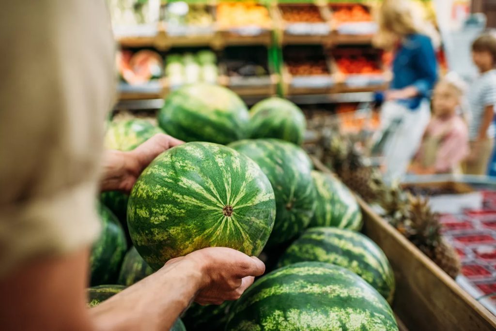 Krupni kadar ruku koje drže i pažljivo pregledaju cijelu, zelenu lubenicu iznad drvenog sanduka punog lubenica u trgovini, dok se u zamućenoj pozadini naziru drugi kupci.