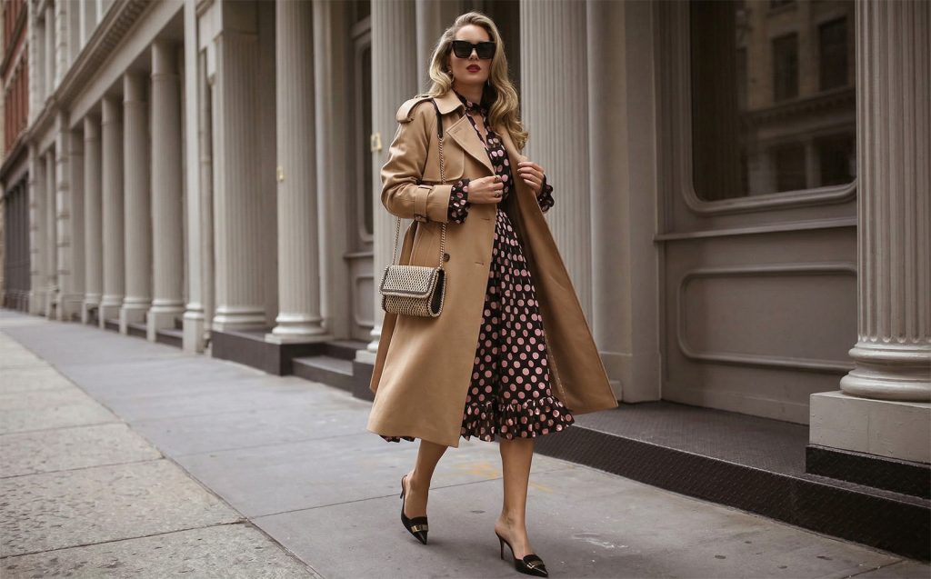 Džaba vam najskuplja odjeća: Ovih 5 sitnica potpuno kvare utisak otmjenosti Žena sa talasastom kosom i naočarima za sunce u hodu na gradskoj ulici. Nosi klasičan bež baloner preko upečatljive crne haljine na pink tufne, uz zlatnu pletenu torbu. Slika zrači samopouzdanjem i vanvremenskim stilom.