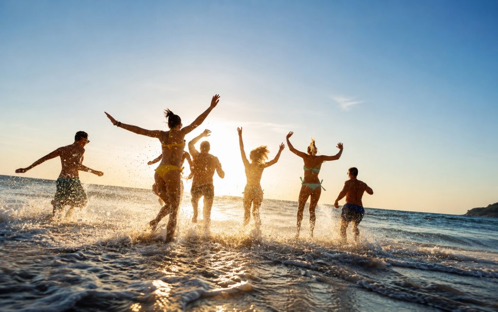 Grupa ljudi trči kroz morske talase na plaži u zalazak sunca, uz atmosferu ljeta, sreće i dobrog raspoloženja.