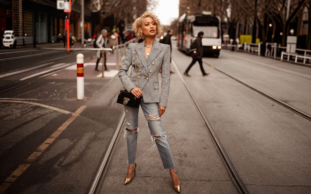 Moderna žena u iscijepanim farmericama, kariranom blejzeru sa zlatnim dugmadima i zlatnim štiklama na gradskoj ulici.