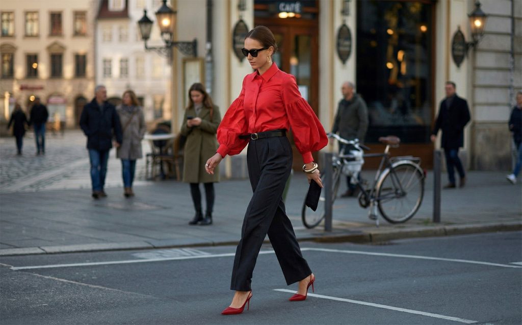 Moderna žena u jarko crvenoj košulji s puf rukavima i crnim hlačama hoda gradskom ulicom.