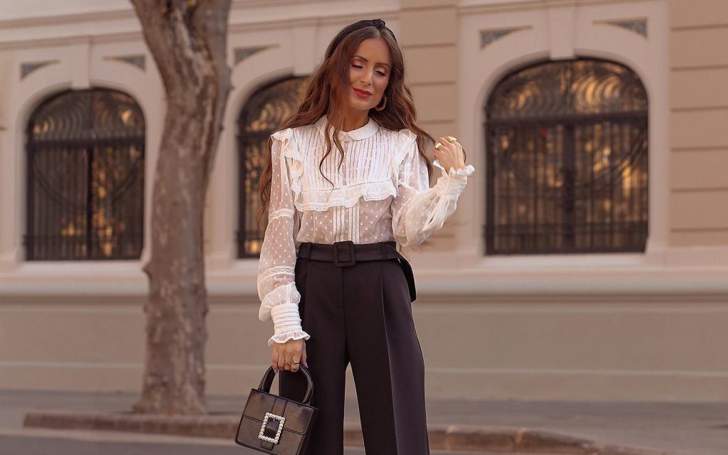 Žena u modernom proljećnom stajlingu koji uključuje bijelu bluzu sa karnerima, tamne hlače visokog struka i crnu torbu sa upečatljivom kopčom.