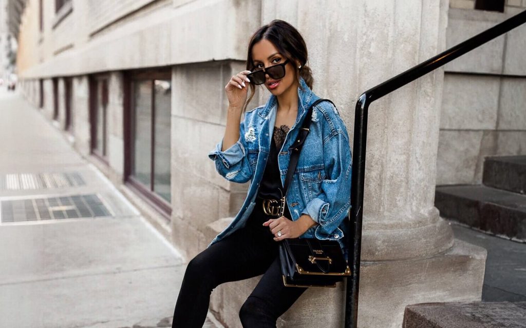 Moderna žena sa sunčanim naočarima pozira napolju noseći plavu teksas jaknu sa ukrasnim dugmadima preko crne majice i hlača.