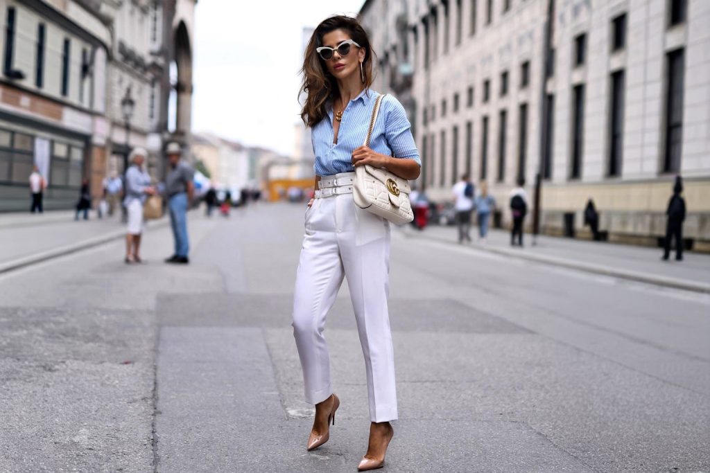 Modna kombinacija za proljeće: žena u svijetloplavoj prugastoj košulji i bijelim hlačama visokog struka sa bež cipelama na petu i bijelom torbom.