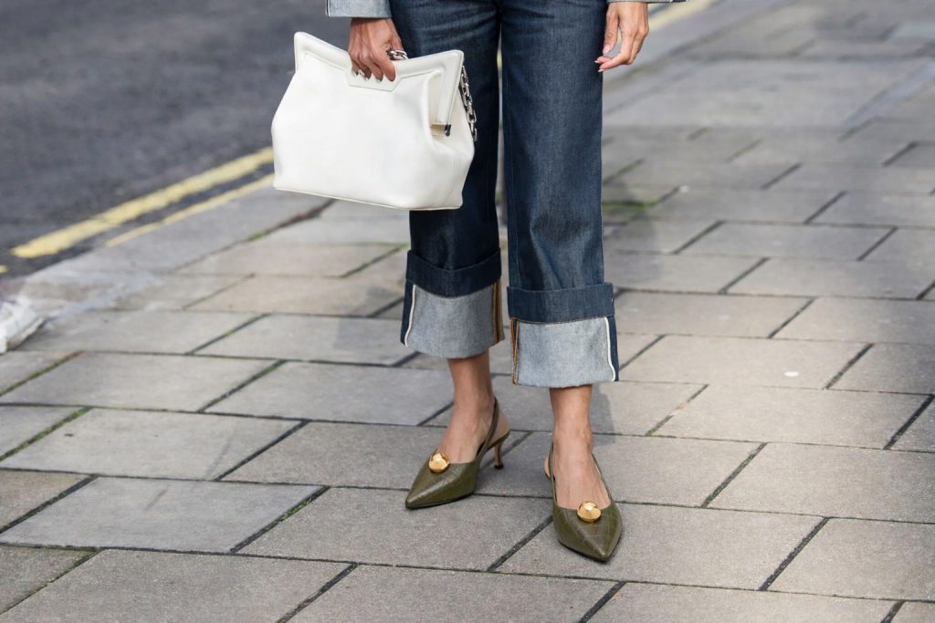 Žena u tamnim farmerkama nosi maslinasto zelene kožne cipele sa zlatnim detaljem i bijelu torbu, ulični stil.