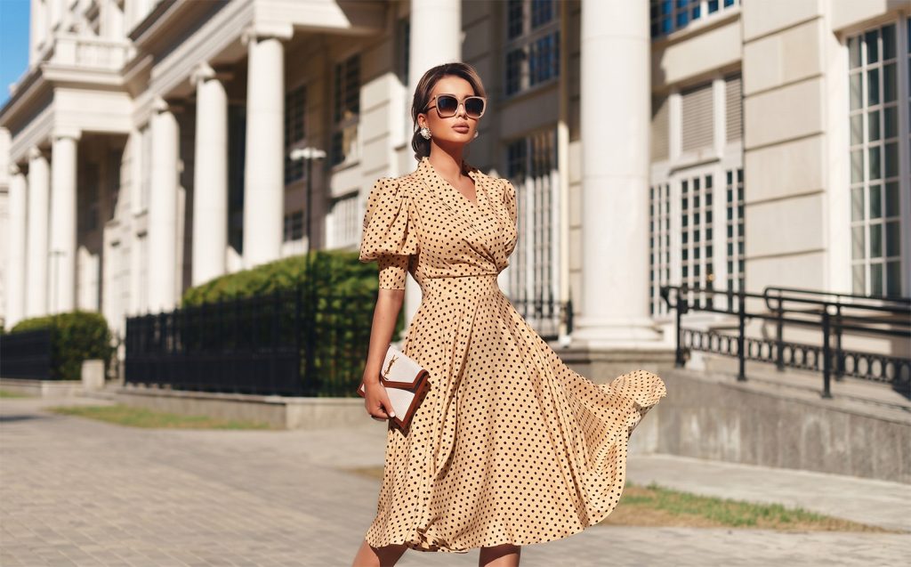 Elegantna žena u bež haljini na crne tačkice sa naglašenim strukom i puf rukavima, hoda ispred zgrade sa bijelim stubovima u modernoj urbanoj sredini.