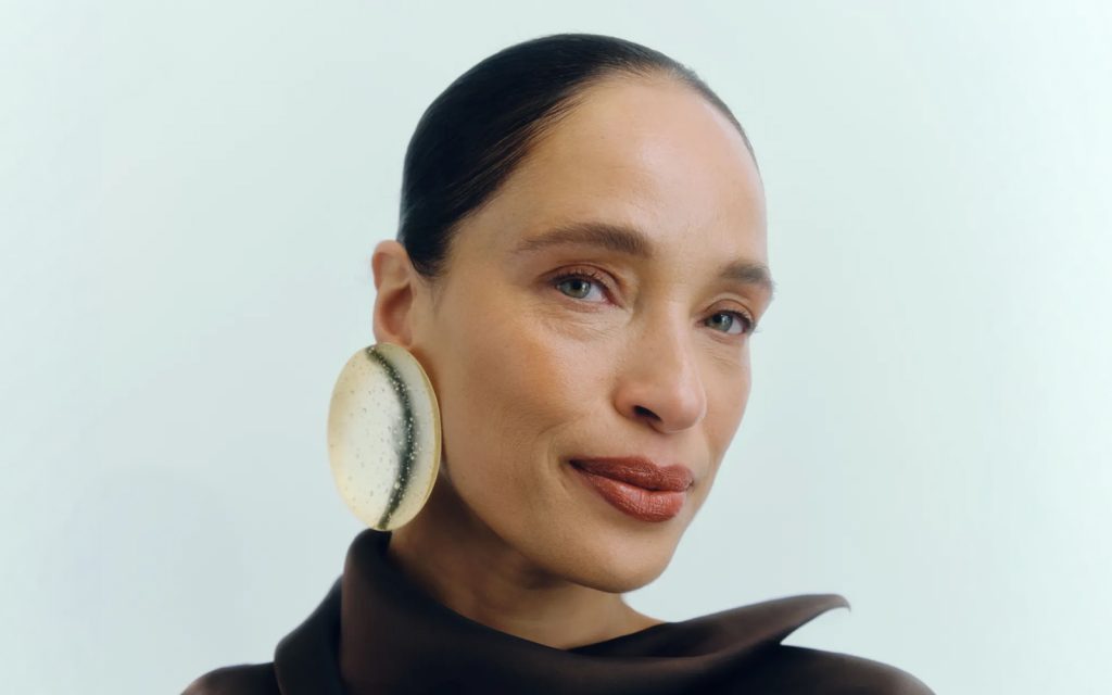 Krupni plan portreta žene sa zalizanom tamnom kosom i zelenim očima. Ona nosi veliku, upečatljivu okruglu minđušu na lijevom uvu. Ima mat smeđi karmin i blagi osmijeh. Pozadina je neutralna svijetloplava.