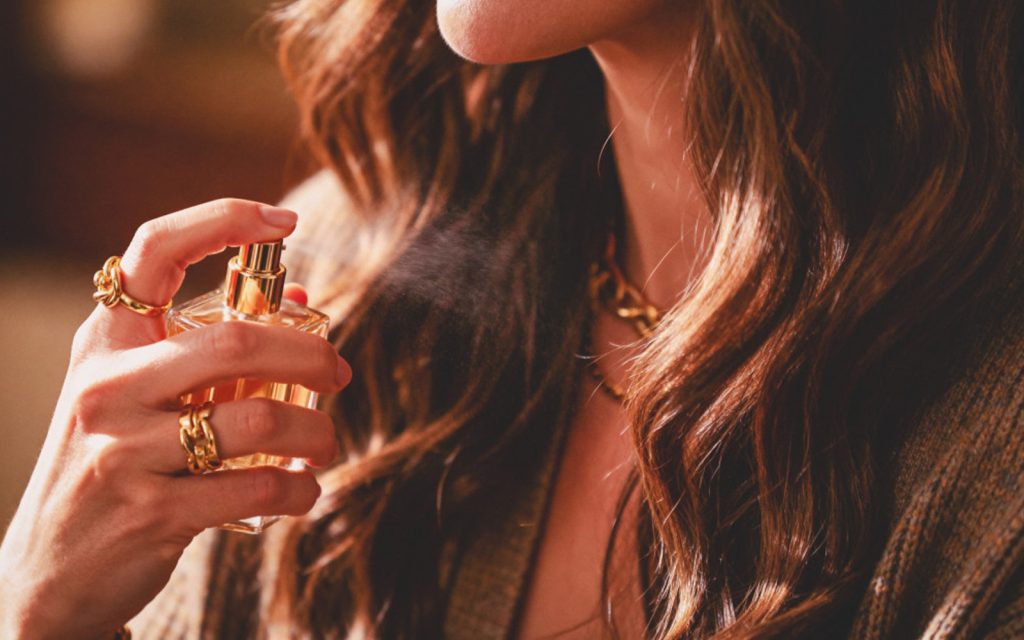 6 mirisnih nota koje dokazano jačaju samopouzdanje Krupni kadar žene sa zlatnim prstenjem koja prska parfem na vrat, toplo osvjetljenje