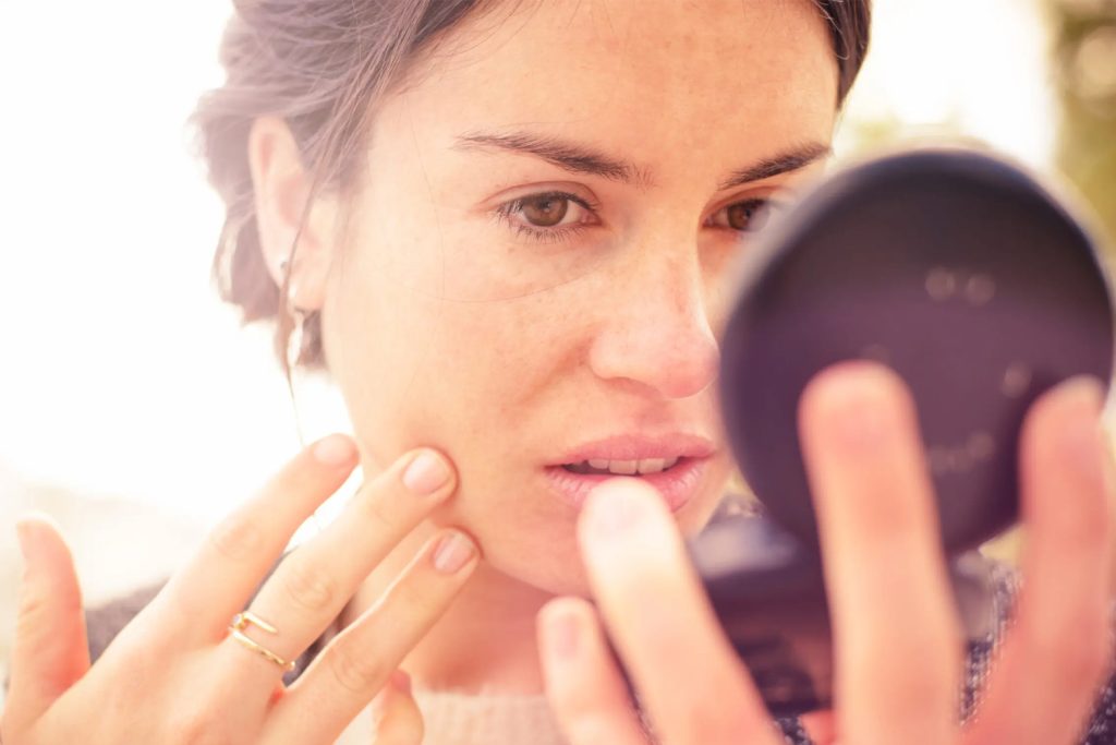 Zabrinuta djevojka u krupnom planu posmatra svoje lice u ogledalu i prstima dodiruje kožu nakon spavanja sa šminkom.