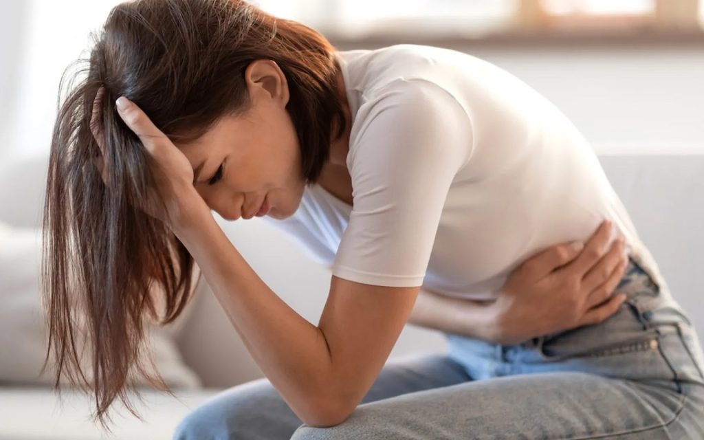 Zdravlje crijeva: Glavni uzrok za stres, anksioznost i umor Mlada žena na kauču drži ruku na glavi i stomaku, s bolnim izrazom lica. Ovo savršeno ilustruje kako problemi sa varenjem mogu dovesti do glavobolje, stresa i anksioznosti, o čemu govori naš članak.