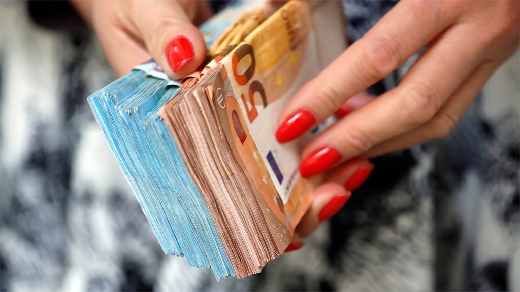 Krupni plan ženskih ruku sa urednim crvenim lakom koje drže debeli svežanj novčanica evra, simbolišući finansijski uspjeh i dobitak.