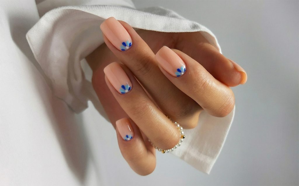 Ženska ruka sa noktima kvadratnog oblika u nježnoj boji breskve, ukrašenim malim plavim cvjetnim detaljima uz korijen nokta.