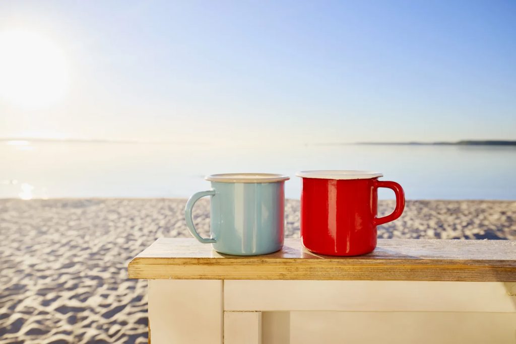 Dvije šolje, svijetlo plava i crvena, na drvenom stolu na pješčanoj plaži obasjane jutarnjim suncem.