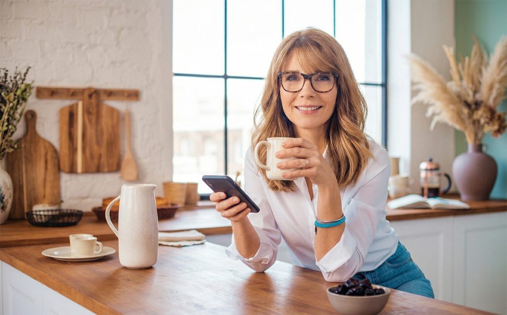 Nasmijana, privlačna žena u pedesetim godinama sa naočalama, u bijeloj košulji, sjedi u svijetloj i modernoj kuhinji, drži šolju u ruci i gleda u kameru, pored zdjele sa suvim šljivama.
