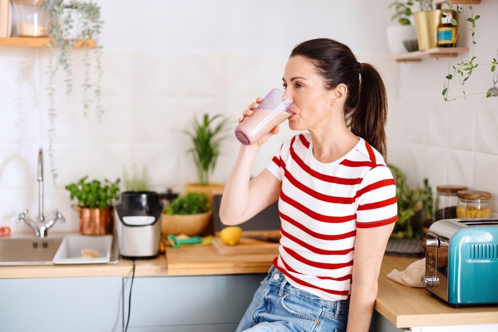 Atraktivna bjelkinja u ranim četrdesetim godinama uživa u zdravom napitku od kolagena u svijetloj i urednoj kuhinji.