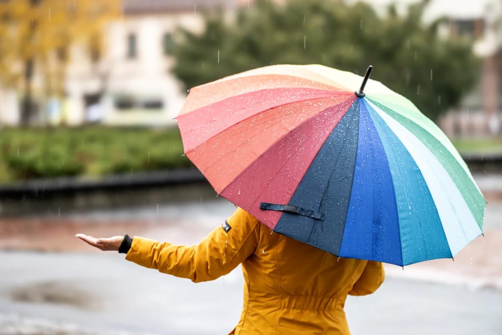 Pogled s leđa na ženu u jarko žutoj kabanici koja drži otvoren šareni kišobran dok hoda po kiši u parku. Jedna ruka joj je ispružena da oseti kapi kiše. Pozadina je blago zamućena, vide se drveće i zgrade.