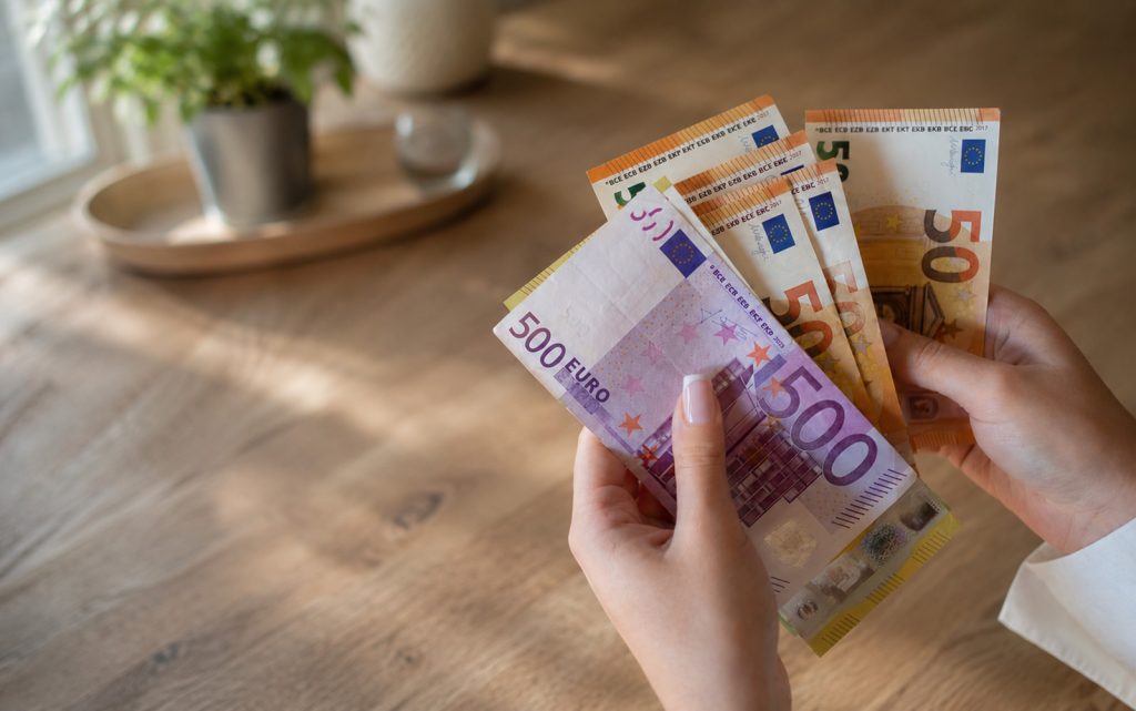 Žena u rukama drži novčanice eura iznad drvenog stola, kao ilustracija finansijskog pomaka u avgustu.