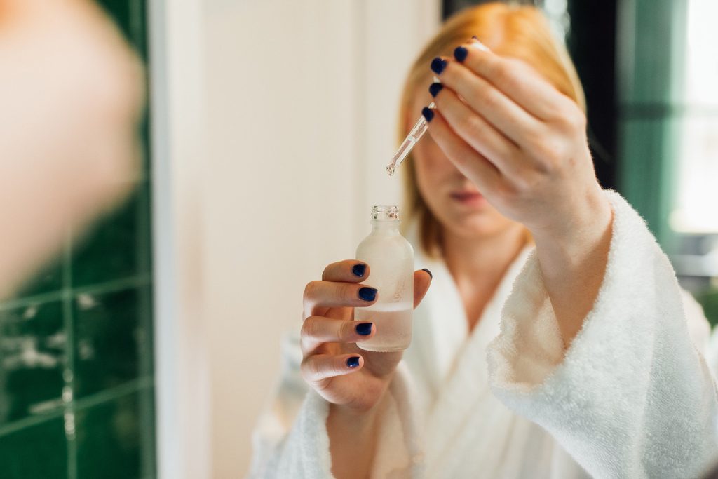 Ceramidi ili peptidi: Šta je bolji izbor za kožu nakon pedesete? Žena u kupatilu drži bočicu seruma s pipetom tokom njege kože nakon pedesete.