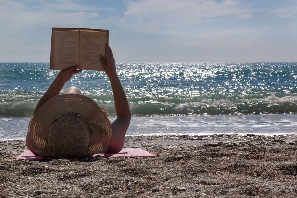 Žena čita knjigu na plaži dok odmara pored mora.